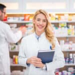 Portrait of a smiling female pharmacist, male colleague working with drugs in the background.