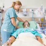 A nurse is standing over a patient laying in a hospital bed, the patient has an oxygen mask on and has other tubes and wires hooked up to him
