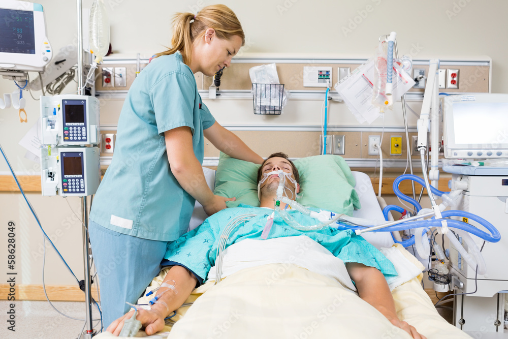 A nurse is standing over a patient laying in a hospital bed, the patient has an oxygen mask on and has other tubes and wires hooked up to him