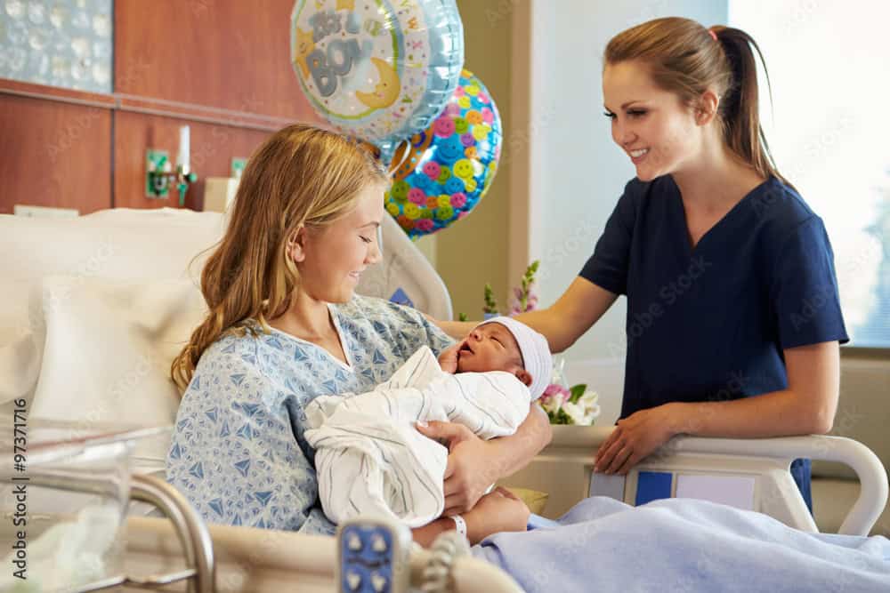 A nurse smiles at a new mother holding her newborn baby in her hospital bed