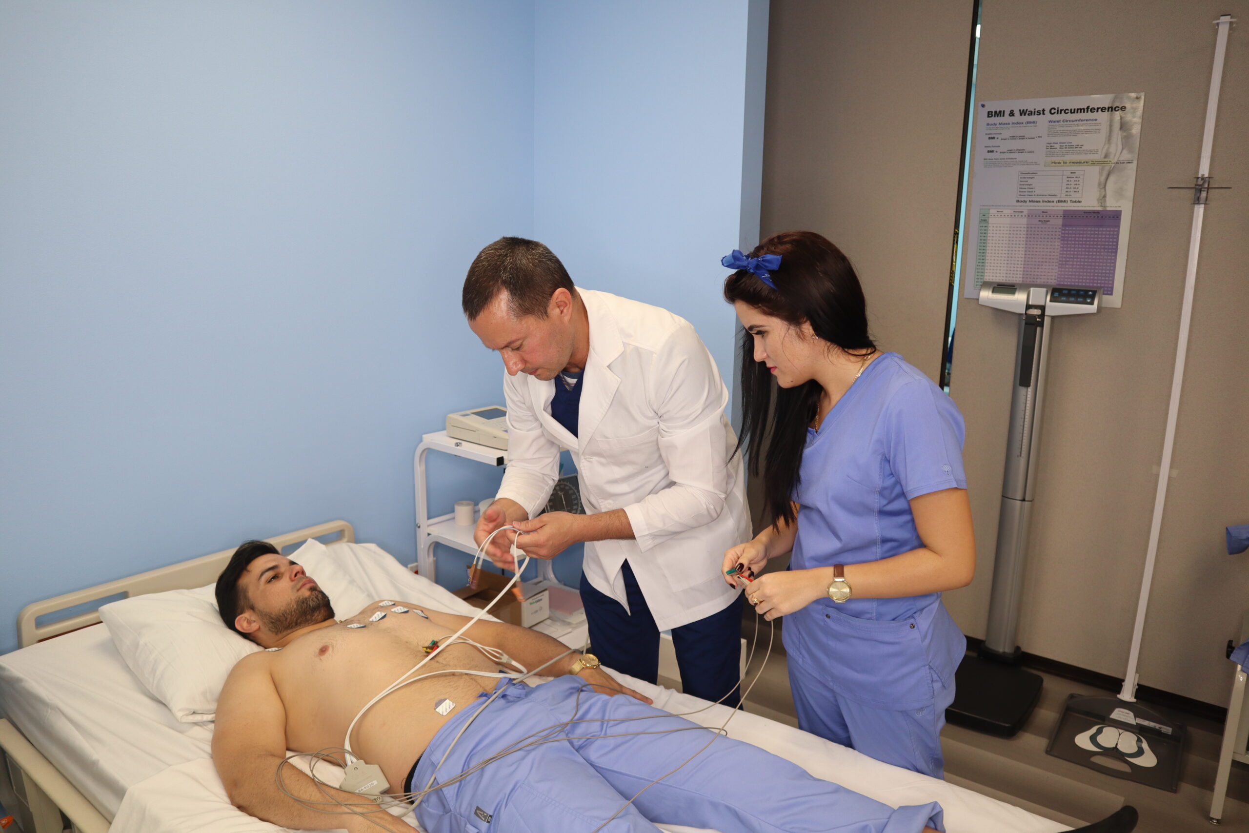 An instructor assists two students who are practicing in a skills lab, one is lying in a hospital bed while the other student stands next to the instructor and observes