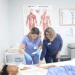 An FVI instructor guides a student practicing hands-on skills with the assistance of another student who is laying in a hospital bed