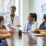 A healthcare professional stands at the end of a conference table talking to a group assembled around the table