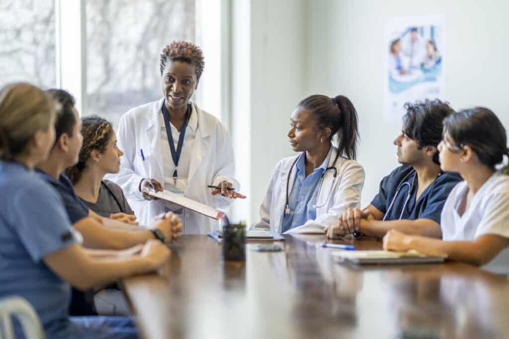 A small group of diverse nursing students sit around a boardroom table as they listen attentively to their teacher and lead doctor.  They are each dressed in medical scrubs and sitting with papers out in front of them.  The doctor is holding out a clipboard with a document on it as she reviews it with the group.