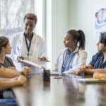 A small group of diverse nursing students sit around a boardroom table as they listen attentively to their teacher and lead doctor. They are each dressed in medical scrubs and sitting with papers out in front of them. The doctor is holding out a clipboard with a document on it as she reviews it with the group.
