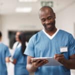 Insurance documents, clipboard and a black doctor writing patient info. Clinic healthcare employee or male nurse with a smile, consulting a medical chart. A man in scrubs, smiling in hospital hallway