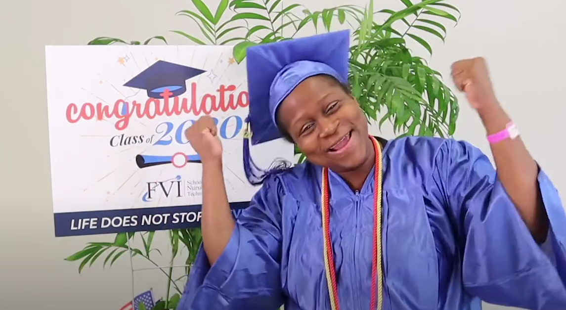An FVI patient care technician celebrates her graduation in her blue cap and gown with a congratulatory sign in the background