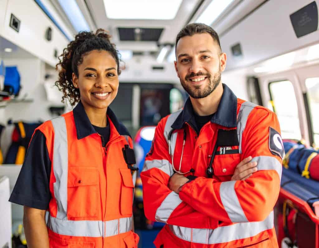 EMT team in bright orange uniforms smiling inside an ambulance.